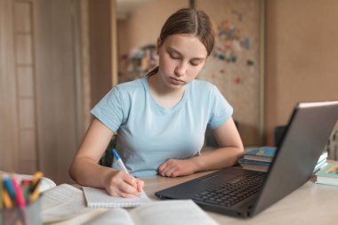 Teen girl does homework, writes lessons in a notebook, notebook monitor, e-learning and online lessons, distance learning. Social networks video call, watching a teacher s lecture video on Internet.