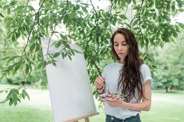Gadis cantik berambut panjang, seniman memegang palet berwarna di tangan, menggambar gambar. Kanvas putih kosong, kulit putih. Musim panas di taman, meninggalkan latar belakang. Emosi kebahagiaan, inspirasi dan kreativitas. — Stok Foto