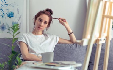 Sanatçı kız evde kanepede oturur, Malbert 'e bakar, resmi inceler, elinde boya fırçası, arka planda çay fincanı olan masa. Duygular, rahatlık, zevk deneyimi, sanat eserlerine hayranlık.