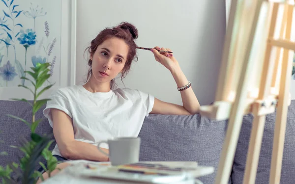 Artist girl sits home sofa, looks malbert, examines painting, holding paintbrush, background table with mug of tea. Emotions inspiration of comfort, pleasure experience, admires work of art.