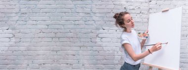 Güzel kadın sanatçı, tuğla duvar arka planı, metin kopyası için boş alan, resim çizer, beyaz tişört, elinde fırça, boya paleti. Beyaz tuval, sehpa