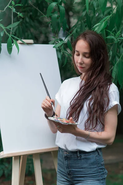 Gadis seniman cantik, di musim panas di taman, menggambar gambar, rambut panjang, kaos putih, kuas di tangan, palet untuk cat. Latar belakang kanvas putih, bingkai, daun pohon. — Stok Foto