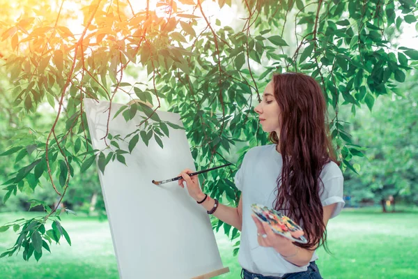 Seniman wanita cantik, di musim panas di taman, menggambar gambar, rambut panjang, kaos putih, kuas di tangan, palet untuk cat. Latar belakang kanvas putih, bingkai, daun pohon. — Stok Foto