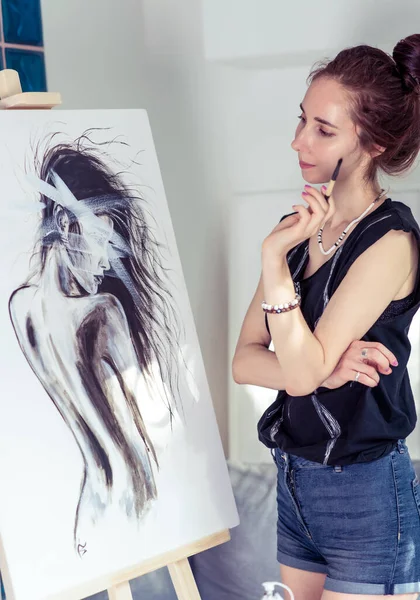 Femme artiste, dessine une silhouette de fille sur une toile blanche, à la maison dans l'atelier, à la main avec un pinceau. Fond, chevalet et mur avec canapé. Regarde le tableau.. — Photo