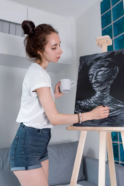 Uma artista, pinta um retrato de uma múmia negra, em casa em uma oficina, em sua mão um pincel, uma caneca com café e chá. Fundo, cavalete e parede com sofá . — Fotografia de Stock