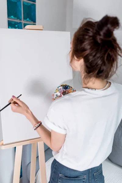 Artystka w białej koszulce, widok z tyłu, w domu w warsztacie, w ręku pędzel i paleta kolorów z farbami. Kanapa w tle, sztalugi i biały obraz. — Zdjęcie stockowe