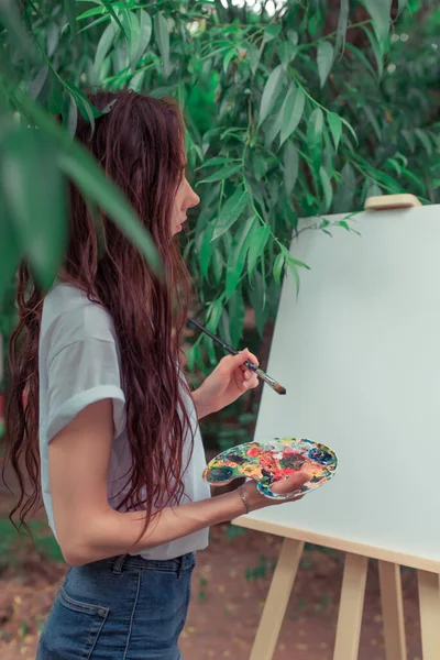 Artis gadis cantik, pemandangan samping, di musim panas di taman, menggambar gambar, rambut panjang, kaus putih, kuas di tangan, palet untuk cat. Latar belakang kanvas putih, bingkai, daun pohon. — Stok Foto