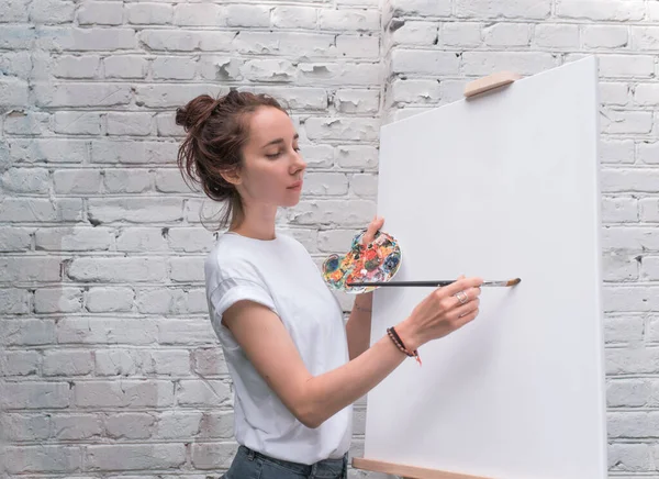 Beautiful woman artist paints a picture, in the summer near a white brick wall, in her hand a brush with paints, the background is an easel. White blank canvas painting. — Stock Photo, Image