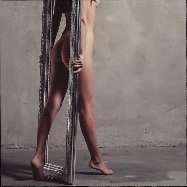 Stüdyoda gri bir duvara karşı çırılçıplak duran genç ve güzel bir kız elinde aynanın boş bir çerçevesini tutuyor.