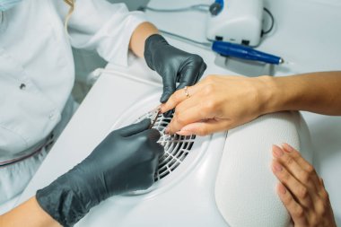 Manikür salonu, usta müşteriye manikür yapıyor. Sabahlık ve maske giymiş bir usta, el çerçevesinde, müşterinin tırnaklarıyla hareket anları.