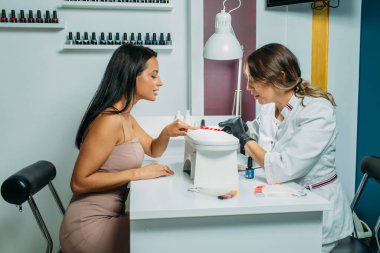 Manikür salonu, usta müşteriye manikür yapıyor. Sabahlık ve maske giymiş bir usta, el çerçevesinde, müşterinin tırnaklarıyla hareket anları.