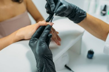 Manikür salonu, usta müşteriye manikür yapıyor. Sabahlık ve maske giymiş bir usta, el çerçevesinde, müşterinin tırnaklarıyla hareket anları.