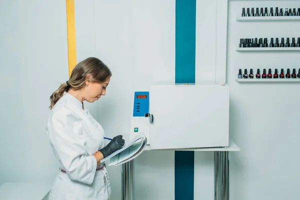 the bright room looks like a medical laboratory, there is an apparatus for quartzing and processing objects in the room. in the frame, a girl in a mask and a medical gown looks very much like a doctor makes an examination and writes in a journal