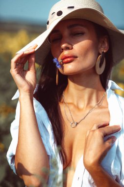 Genç ve güzel bir kız, anlamlı bir yüzü ve güzel dudakları var, parlak sarı bir ayçiçeği tarlasının ortasında duruyor, güzel bir şapkası ve çıplak vücudunda beyaz bir gömleği var.