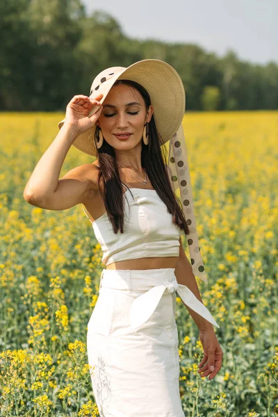 A young beautiful girl with a good figure, stands among a spacious field with yellow flowers, she is dressed in summer light clothes and wears a beautiful hat, summer, sun and bright colors