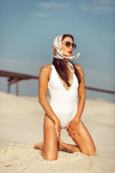 A young beautiful girl with a good figure, around only white sand and blue sky, and on her a white swimsuit and beautiful accessories