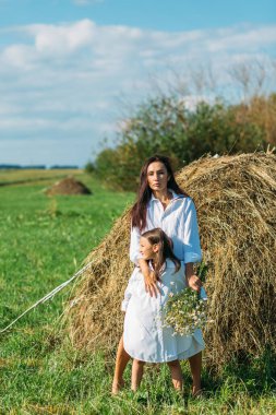 Anne ve kız, beyaz gömlekler içinde ve çiçekli, yazın tarlalarda yürüyorlar. Güneş, yeşillik, tarla, çimen, saman, gökyüzü.