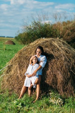 Anne ve kız, beyaz gömlekler içinde ve çiçekli, yazın tarlalarda yürüyorlar. Güneş, yeşillik, tarla, çimen, saman, gökyüzü.