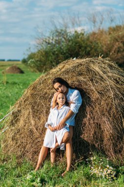 Anne ve kız, beyaz gömlekler içinde ve çiçekli, yazın tarlalarda yürüyorlar. Güneş, yeşillik, tarla, çimen, saman, gökyüzü.