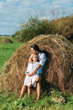 Anne ve kız, beyaz gömlekler içinde ve çiçekli, yazın tarlalarda yürüyorlar. Güneş, yeşillik, tarla, çimen, saman, gökyüzü.