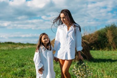 Anne ve kız, beyaz gömlekler içinde ve çiçekli, yazın tarlalarda yürüyorlar. Güneş, yeşillik, tarla, çimen, saman, gökyüzü.