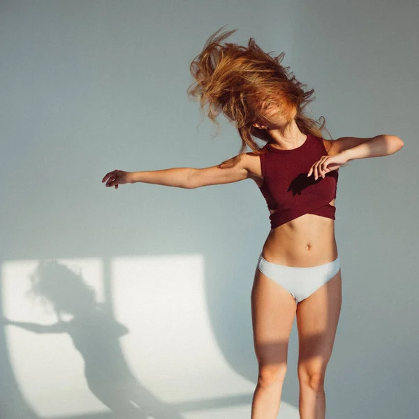 A young girl, with a plastic figure, jumps on a white background in the studio, the sunlight from the window shines on her and creates a picture on the floor. she\'s wearing a sports top and shorts