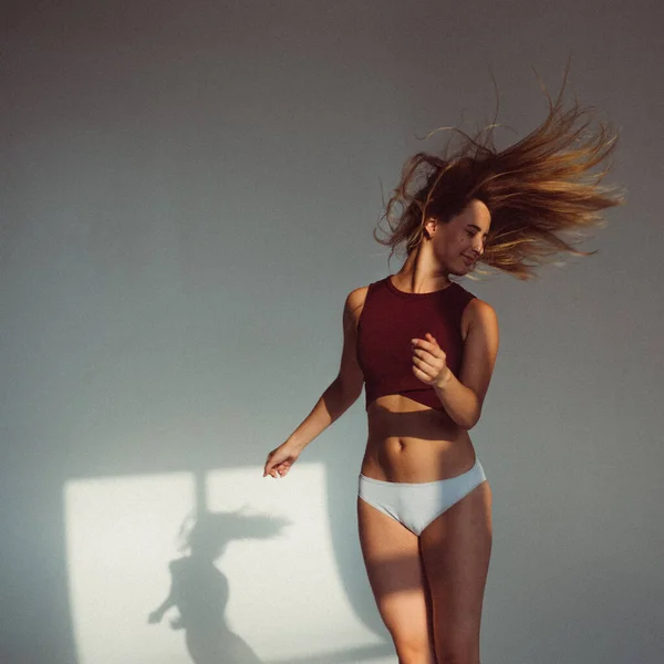 A young girl, with a plastic figure, jumps on a white background in the studio, the sunlight from the window shines on her and creates a picture on the floor. she\'s wearing a sports top and shorts