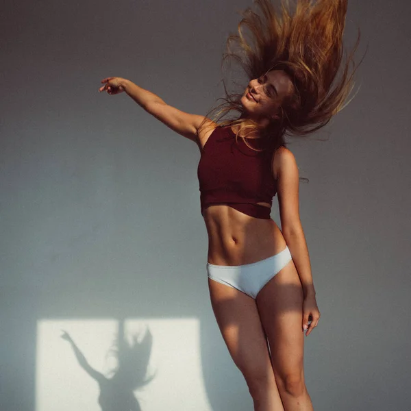 A young girl, with a plastic figure, jumps on a white background in the studio, the sunlight from the window shines on her and creates a picture on the floor. she\'s wearing a sports top and shorts