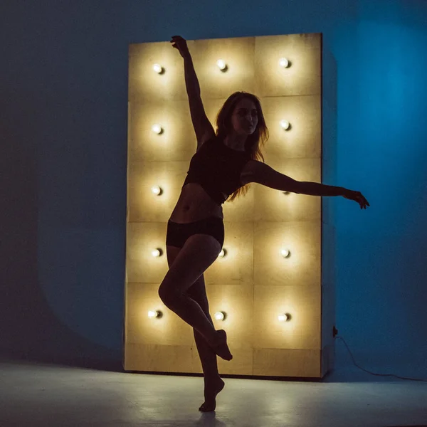 A young beautiful girl, with a plastic figure, makes dance elements near the wall with burning yellow lamps, the light illuminates the contours of her figure. she\'s wearing a sports top and shorts