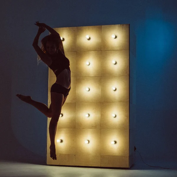 A young beautiful girl, with a plastic figure, makes dance elements near the wall with burning yellow lamps, the light illuminates the contours of her figure. she\'s wearing a sports top and shorts