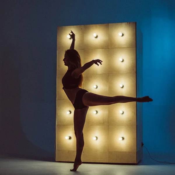 A young beautiful girl, with a plastic figure, makes dance elements near the wall with burning yellow lamps, the light illuminates the contours of her figure. she\'s wearing a sports top and shorts