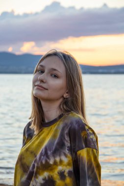 Tatlı tatlı gülümseyen genç bir kız bulutlu bir yaz manzarası, sakin bir deniz ve arkasında güneşin kaybolduğu dağlarla kumlu bir sahilde duruyor.