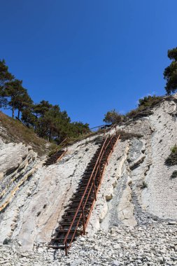 Vahşi bir sahilde eski metal bir merdiven ve büyük beyaz kayalar. Kamp alanına giden yol ormandan geçiyor. Açık bir günde yaz manzarası. Rusya 'nın Gelendzhik tatil beldesinin çevresi