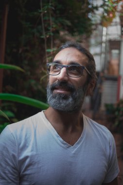 Bahçede gülümserken, uzun, gri saçlı ve yana bakan gözlüklü bir adamın portresinin dikey fotoğrafı.