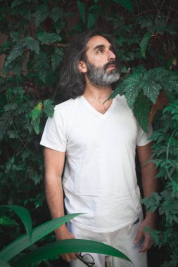 Bahçenin ortasında beyaz giyinmiş, uzun gri saçlı bir adamın düşünceli bir ifadeyle bitkileri düşünürken çekilmiş dikey fotoğrafı.