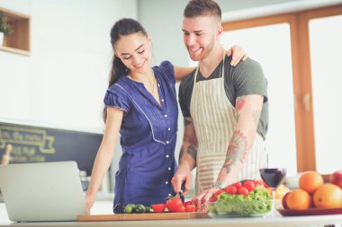 Sebze doğrayan genç bir adam ve mutfakta dizüstü bilgisayarı olan bir kadın.