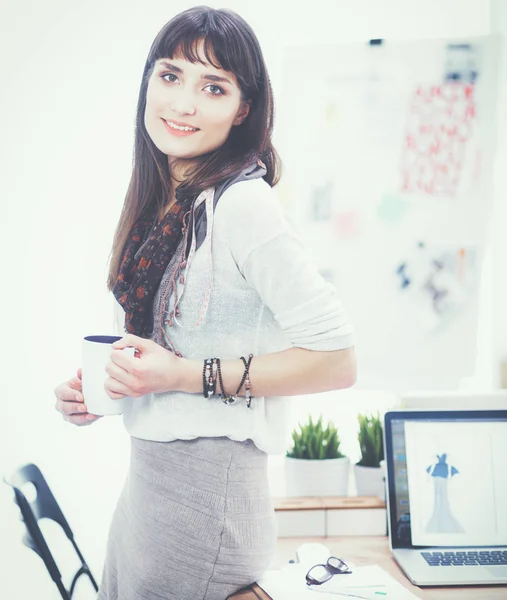 Junge attraktive Modedesignerin steht im Büro am Schreibtisch und hält Tasse — Stockfoto