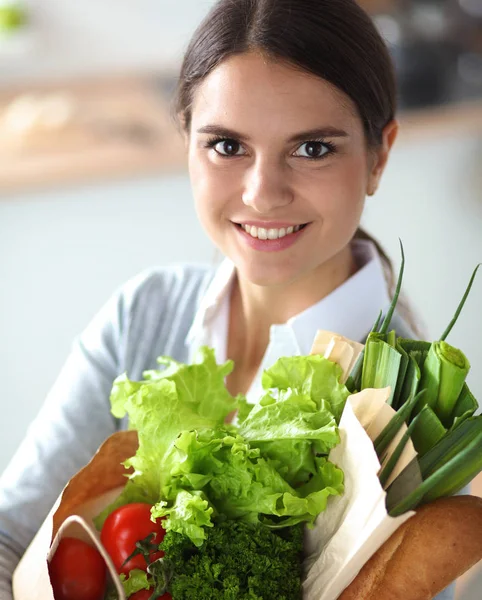 Mujer joven sosteniendo bolsa de la compra de comestibles con verduras. De pie en la cocina — Foto de Stock