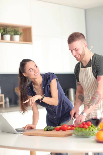 Junger Mann schneidet Gemüse und Frau steht mit Laptop in der Küche — Stockfoto