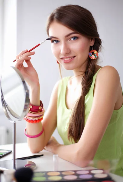 Joven hermosa mujer haciendo maquillaje cerca del espejo, sentado en el escritorio — Foto de Stock