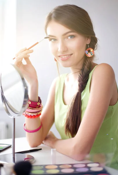 Joven hermosa mujer haciendo maquillaje cerca del espejo, sentado en el escritorio —  Fotos de Stock