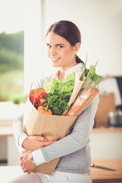 Молода жінка тримає продуктовий сумку з овочами. Стоячи на кухні — стокове фото
