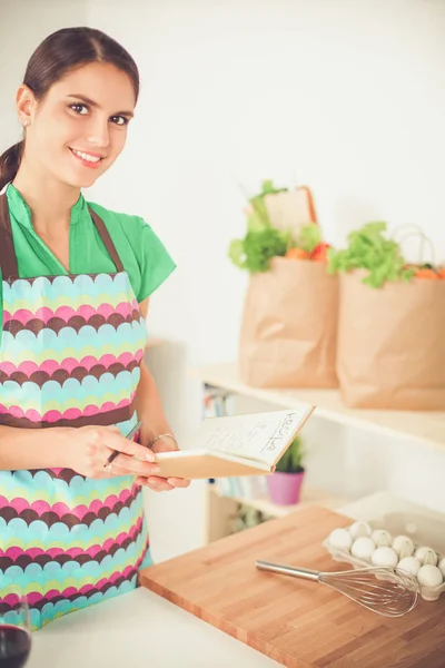 Happy beautiful woman standing in her kitchen writing on a notebook at home — Stock Photo, Image