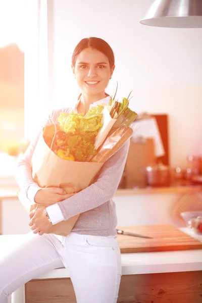 Mujer joven sosteniendo bolsa de la compra de comestibles con verduras. De pie en la cocina — Foto de Stock