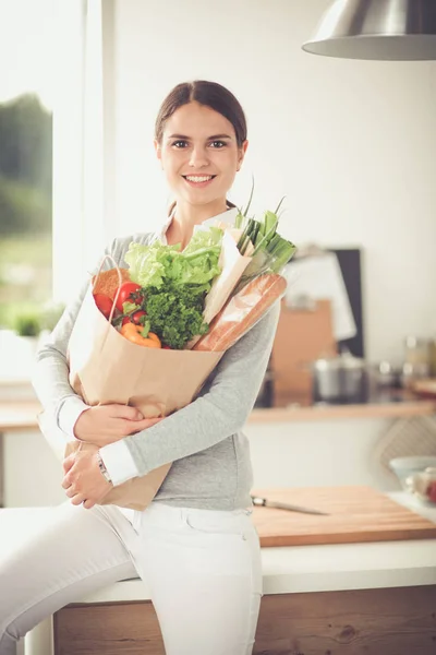 Mujer joven sosteniendo bolsa de la compra de comestibles con verduras. De pie en la cocina — Foto de Stock