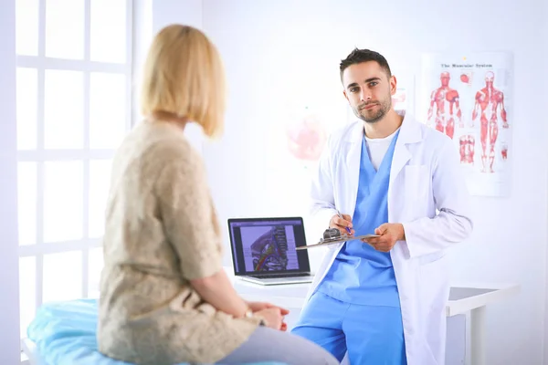 Handsome doctor is talking with young female patient and making notes while sitting in his office — Stock Photo, Image