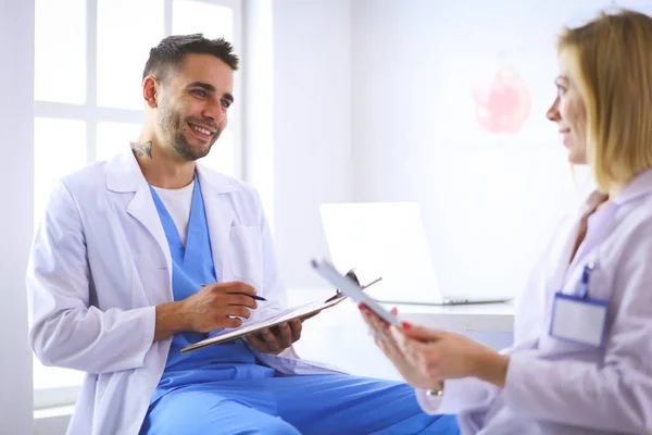 Knappe dokter praat met jonge vrouwelijke patiënt en maakt aantekeningen terwijl hij in zijn kantoor zit — Stockfoto