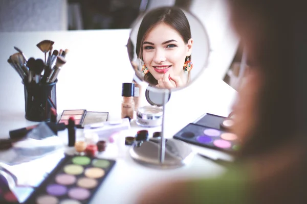 Joven hermosa mujer haciendo maquillaje cerca del espejo, sentado en el escritorio —  Fotos de Stock