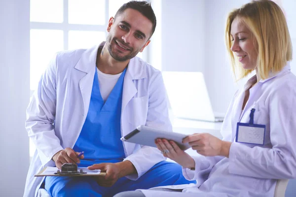 Knappe dokter praat met jonge vrouwelijke patiënt en maakt aantekeningen terwijl hij in zijn kantoor zit — Stockfoto