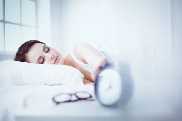 Rahat yatakta yatarken ve blissfully çalar saat arka plan üzerinde uyuyan güzel genç kadın — Stok fotoğraf